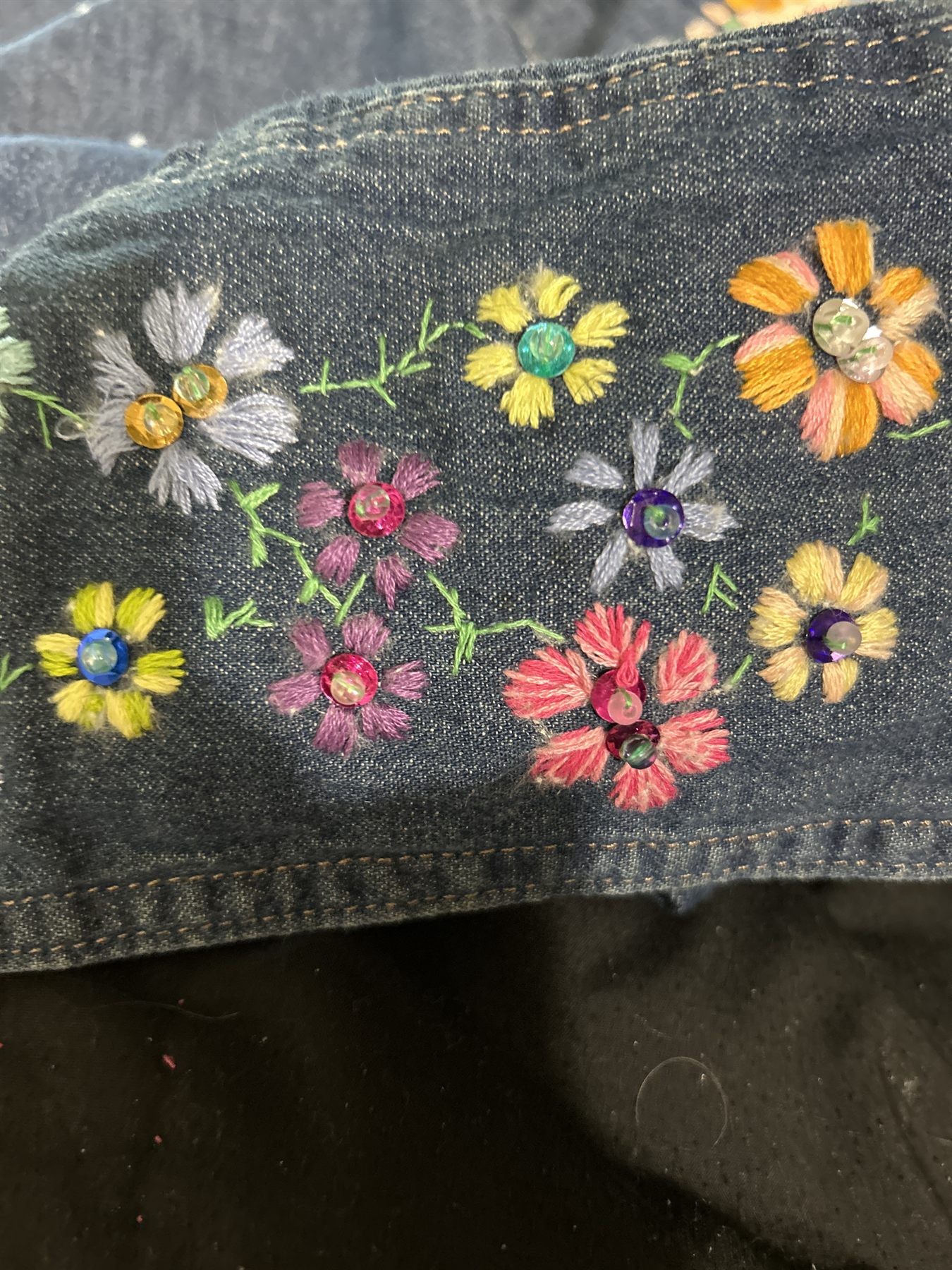 Worn denim covered in small hand-embroidered flowers in yellow, coral, lilac and orange, each with a sequin or rhinestone center.