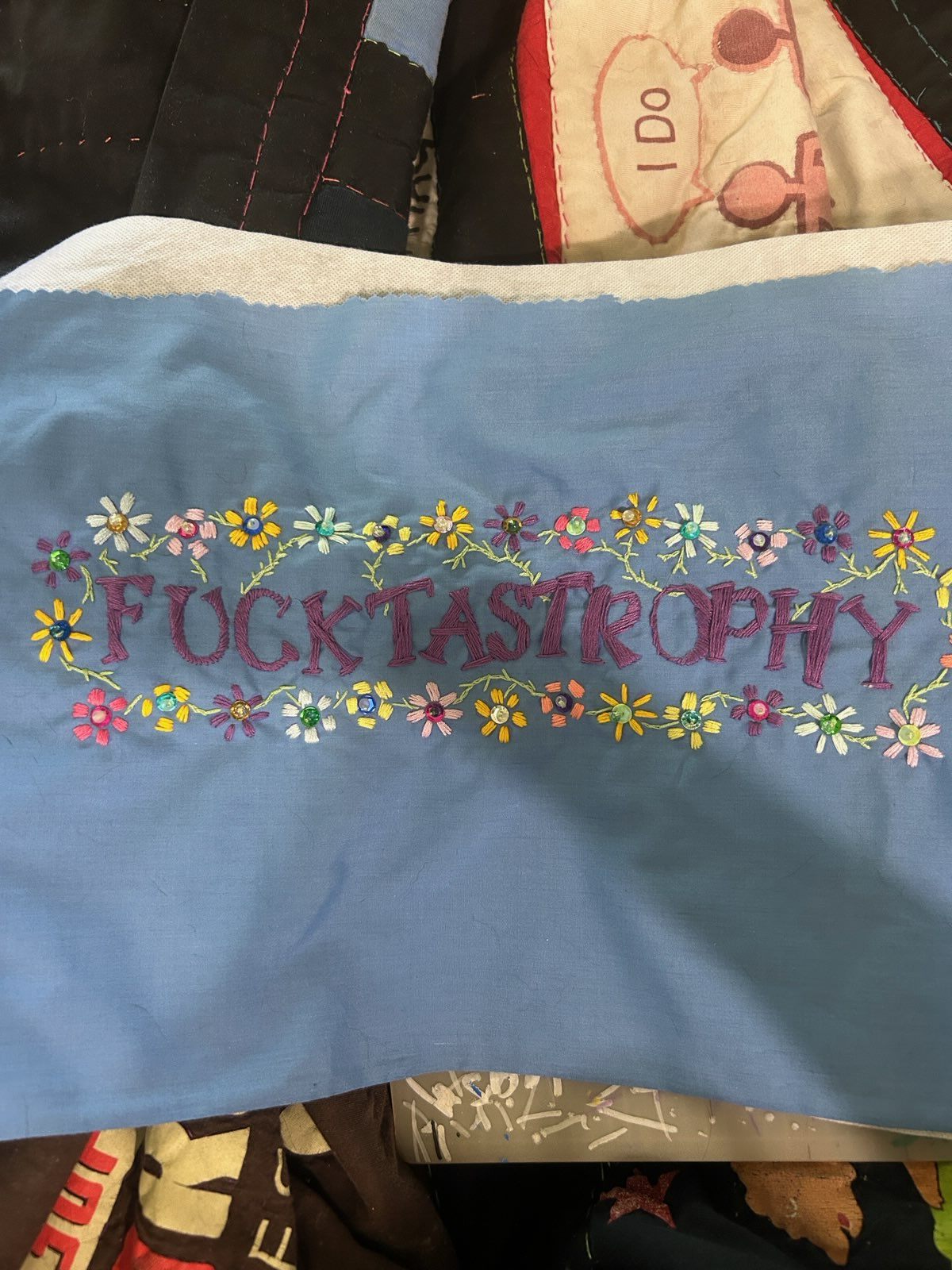 Blue cotton panel embroidered with the word Fucktastrophy in mulberry, framed above and below by rows of small flowers with rhinestone centers.